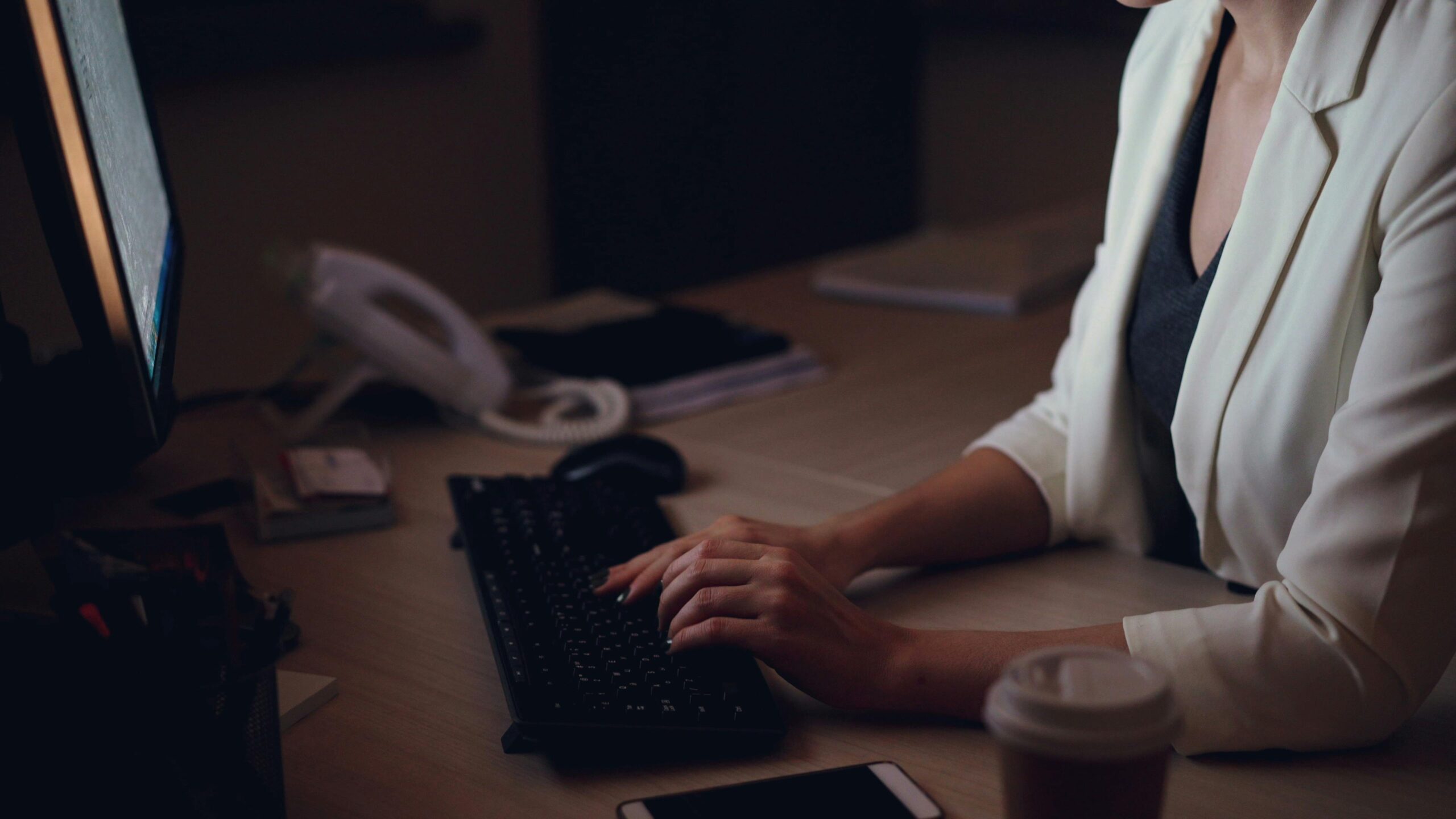 Woman in white blazer typing on keyboard late at night in office.